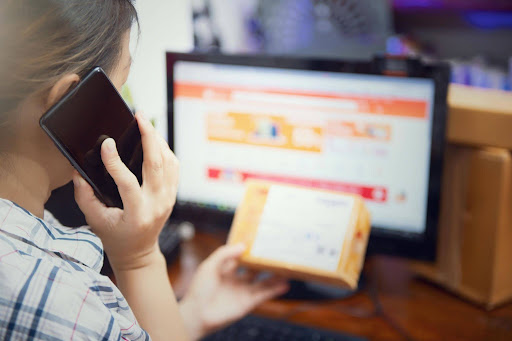 A business owner talking on a smartphone while holding a small shipping package in front of a computer screen showing an e-commerce dashboard.