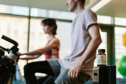 Two gym-goers working out on stationary bikes in a bright fitness center with a bottle of dietary softgels in the foreground, showcasing a product from an EPROLO POD supplement supplier.