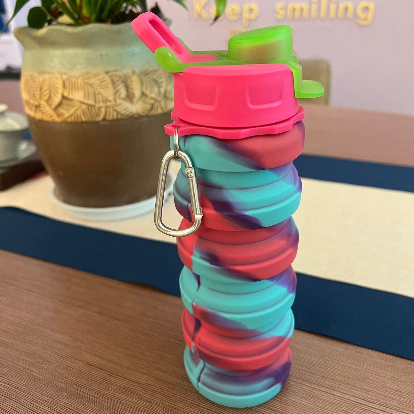 A colorful, tie-dye collapsible silicone water bottle with an attached metal carabiner clip resting on a wooden table, highlighting one of the best EPROLO summer dropshipping products.