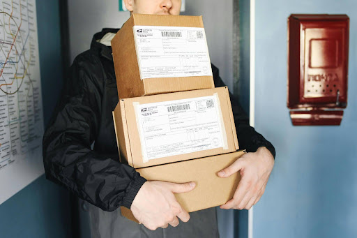 A delivery courier carrying a high stack of labeled cardboard shipping boxes near a mailbox, illustrating the fulfillment and shipping process for EPROLO private label supplements dropshipping.