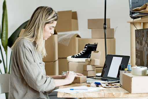 A focused business owner working on her laptop while preparing a small package at a desk surrounded by shipping boxes, highlighting the dedication required for an effective EPROLO repeat customer strategy.