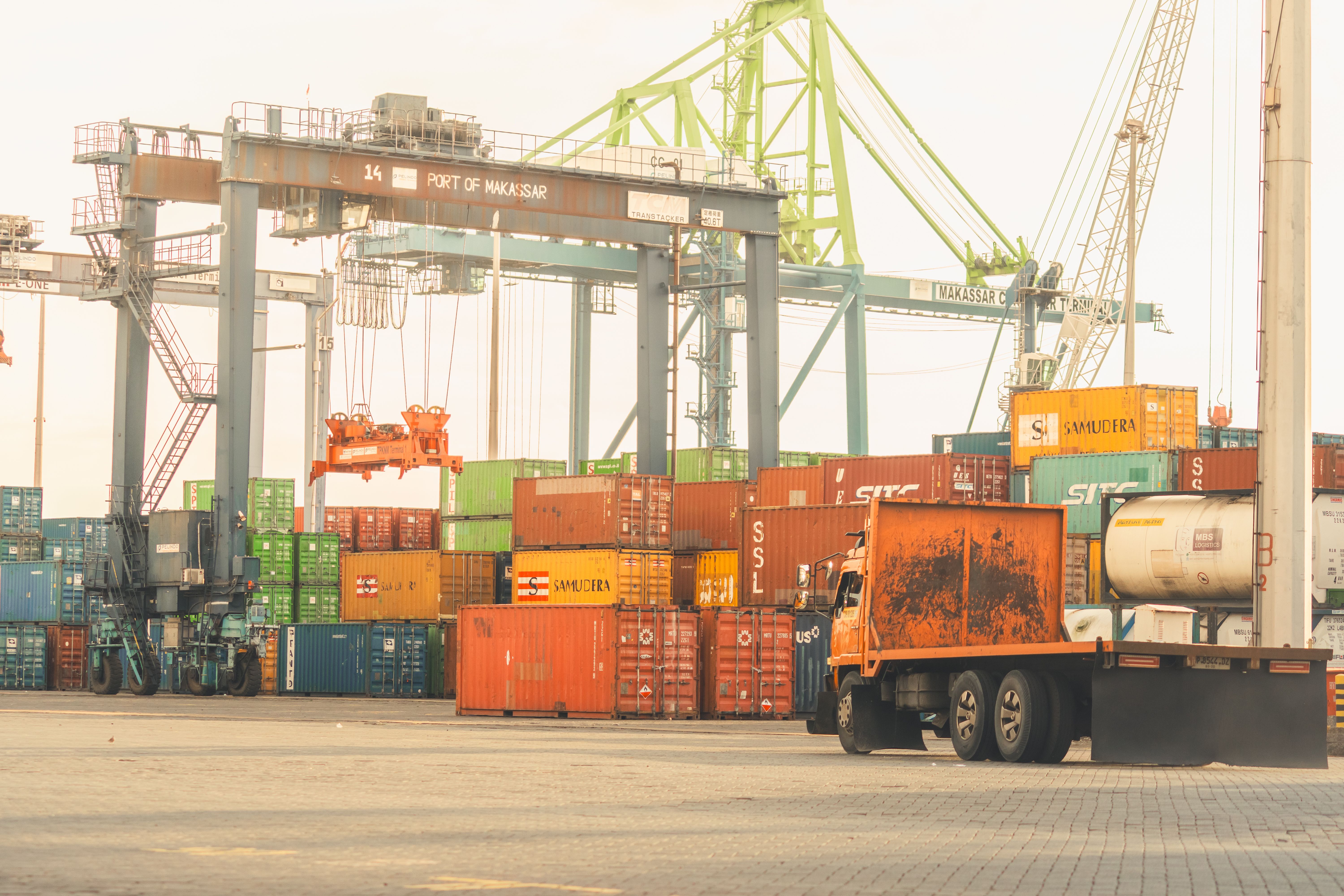A massive gantry crane organizing shipping containers near a flatbed cargo truck at a bustling port, illustrating the global logistics and clearance infrastructure involved in EPROLO EU customs duty 2026.