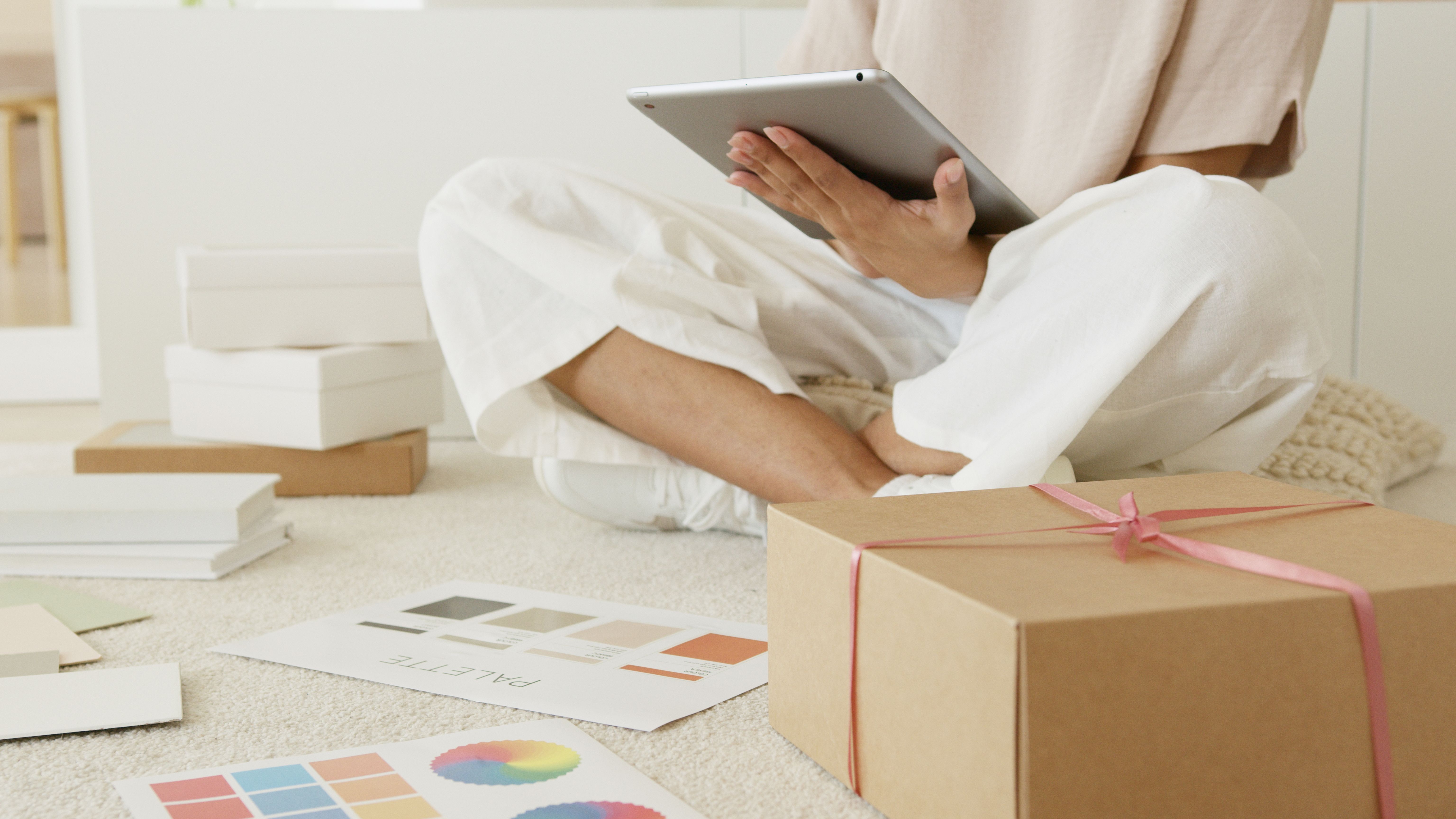 A merchant sitting cross-legged on the floor reviewing cross-border compliance on a tablet alongside design palettes and shipping boxes, illustrating the guidelines within the EPROLO merchant playbook customs duty.