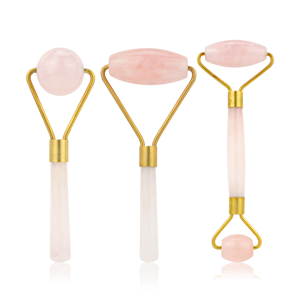 Three rose quartz facial massage tools: spherical roller, flat stone, and double‑ended roller with brass accents on cream handles
