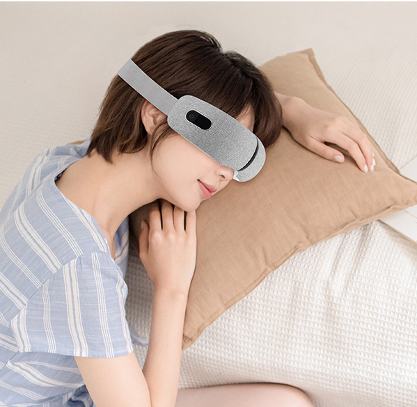 Woman relaxing in bed wearing the eye massager, eyes closed, soft lighting
