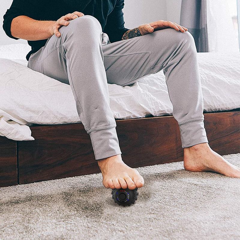 Man in casual gray clothing using massage ball on foot while sitting on bed in a sunlit home setting