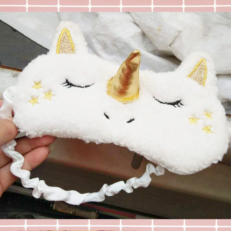 Hand holding white plush unicorn sleep mask with gold metallic horn, glitter gold ears, embroidered gold stars and eyelashes, and white satin ruched elastic band