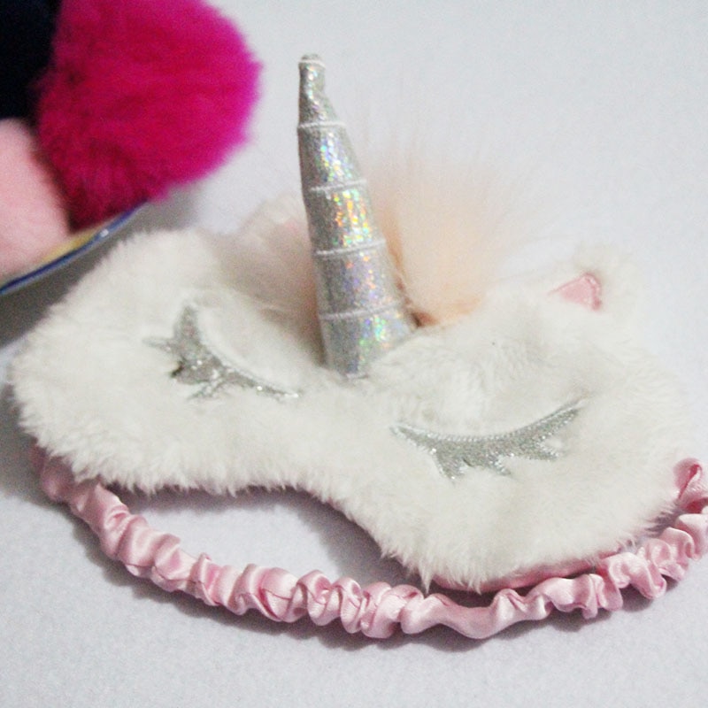 Close-up of white plush unicorn sleep mask with holographic silver spiral horn, feather accent, glitter star eyelashes, and pink satin elastic band