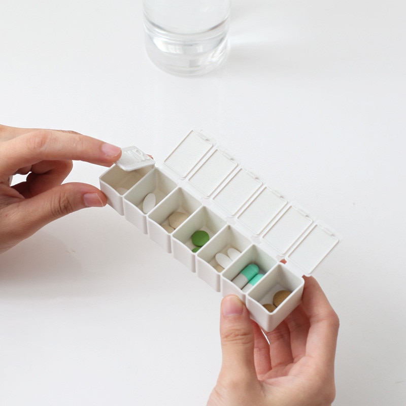Hands opening white pill organizer filled with assorted medications next to a glass of water