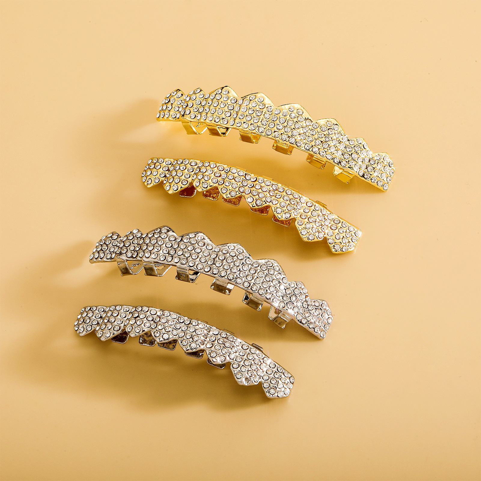 Four iced-out tooth grillz pieces laid flat on warm beige background showing two gold and two silver rhinestone-paved sets